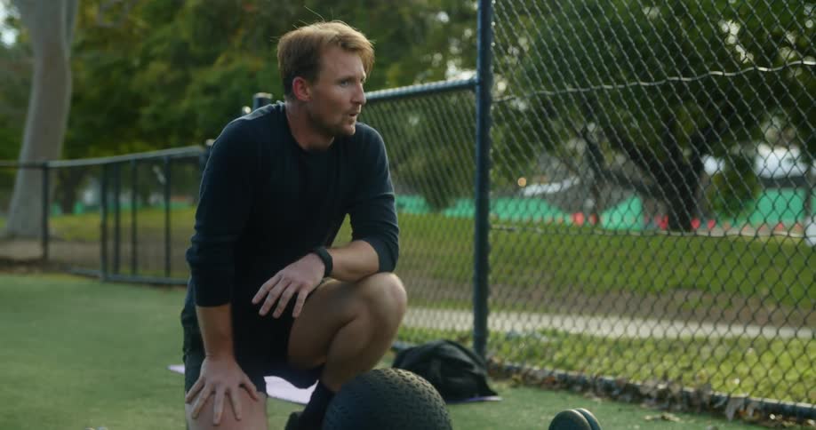 A man in a public park breathes heavily after exercising and looks at his watch