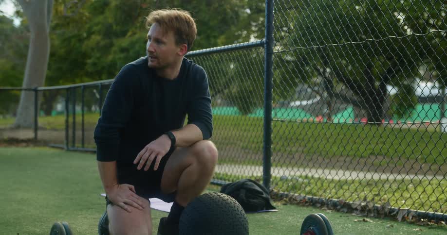 A man kneels on the ground and counts while checking his watch as he takes a break from exercise in a park
