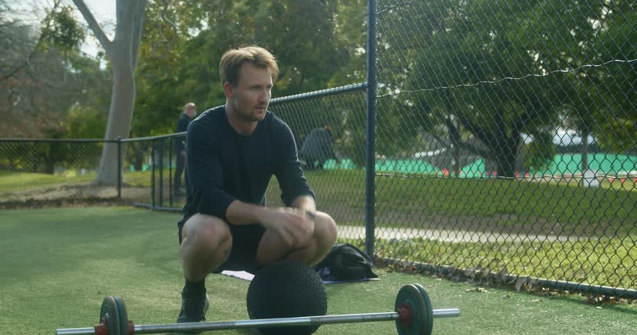 A middle aged man checks his watch while squatting in front of a barbell in an outdoor exercising area