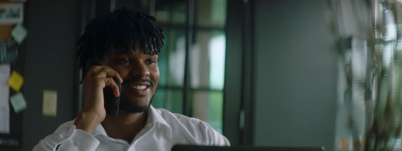 Black man with braid hair a beard and a white shirt sits at a table and talks on the smartphone
