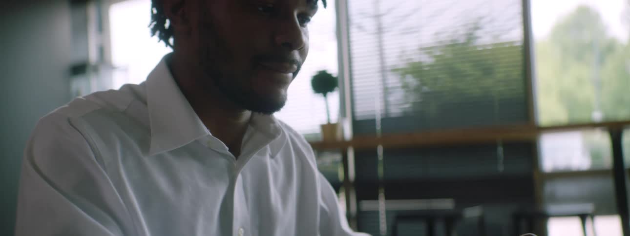A man sits at a table and typing on a laptop with his cell phone beside him