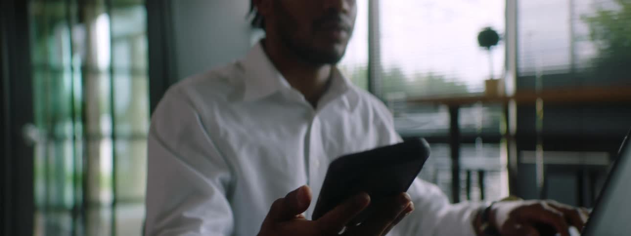 A young man pauses from typing on his laptop in order to answer a call on his cell phone