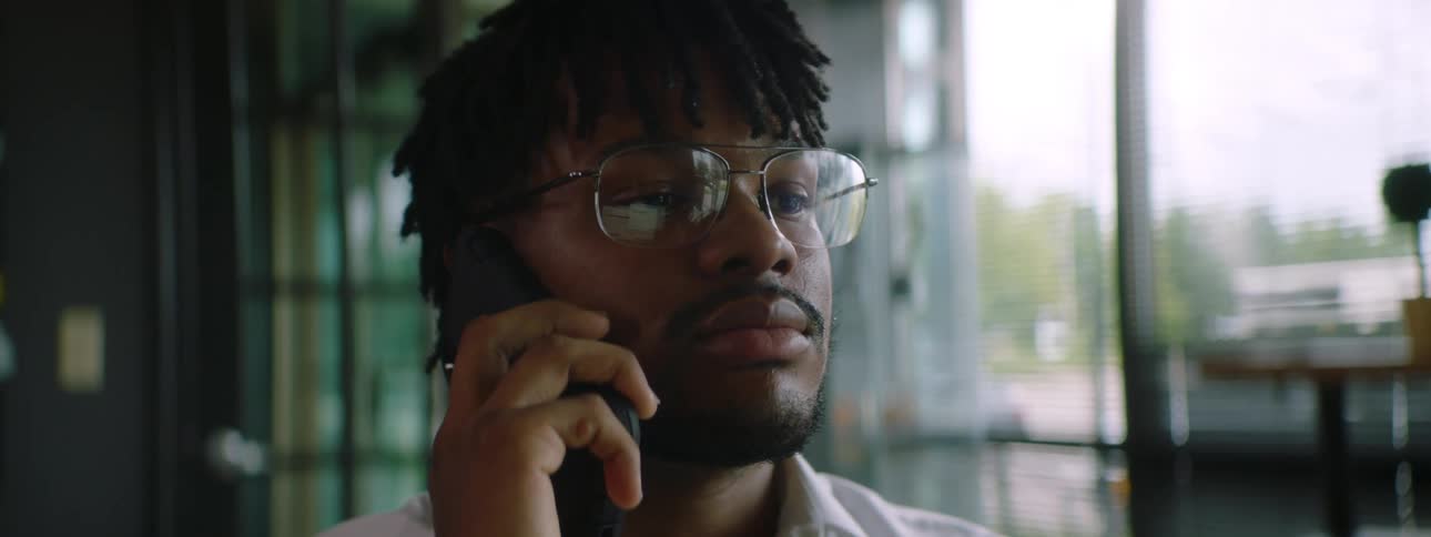 Black man with braided hair glasses and a beard talks on the smartphone in a room