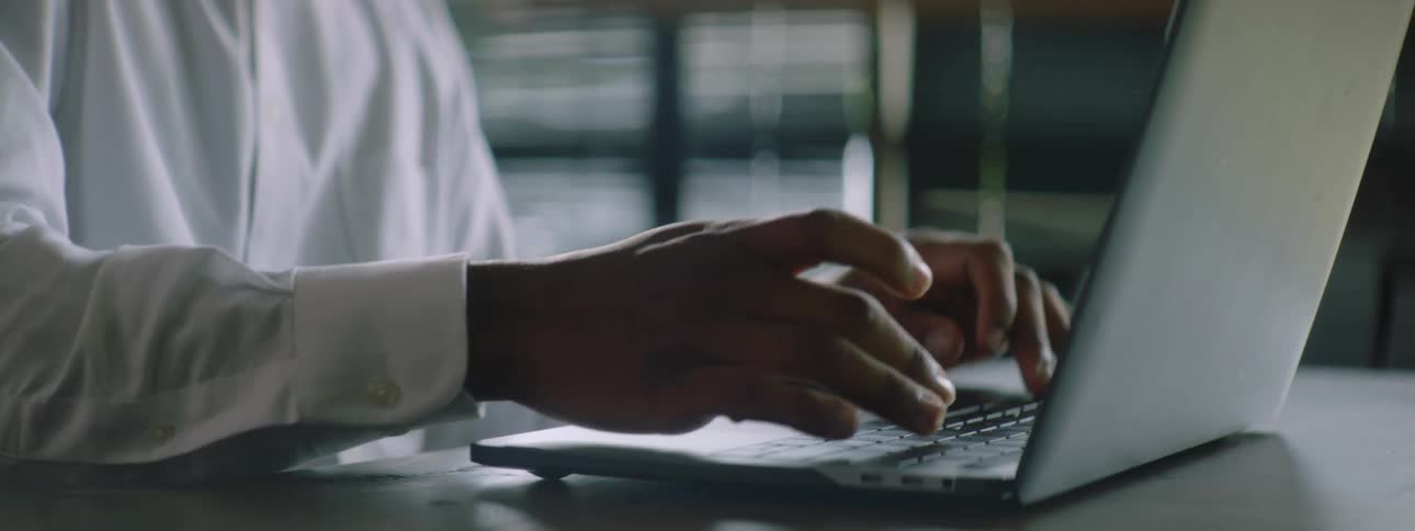 Black man with braided hair a beard and a white shirt uses a grey laptop while talking on a smartphone