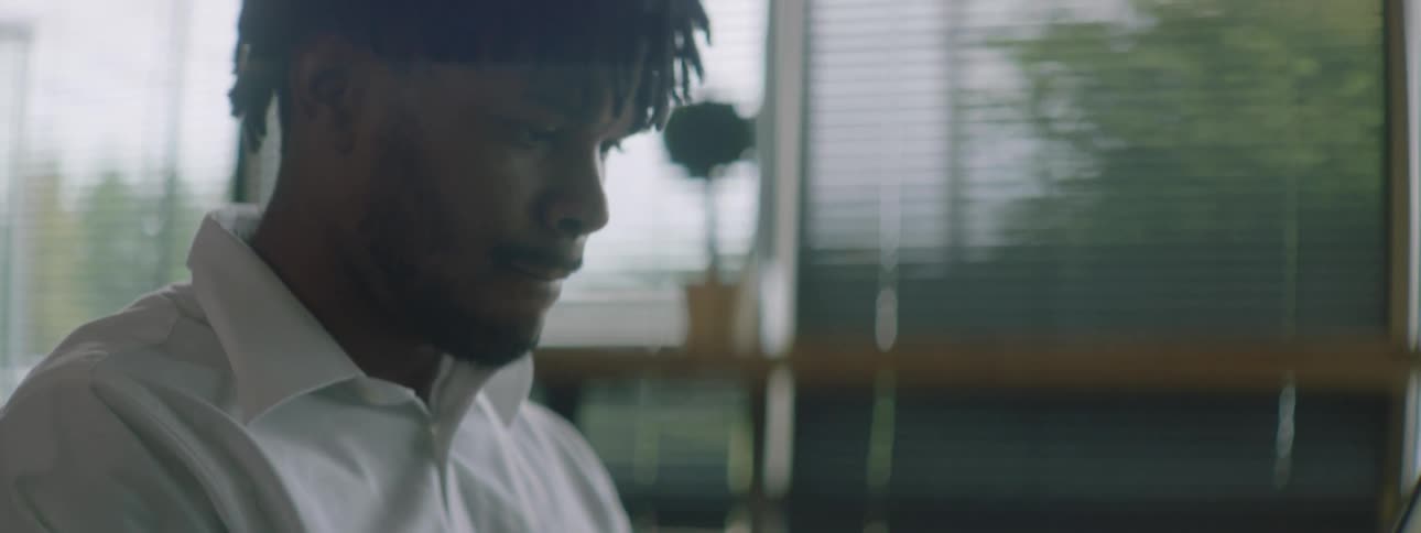 Black man with braids a beard and a white shirt sitting at a table uses a grey laptop in a room