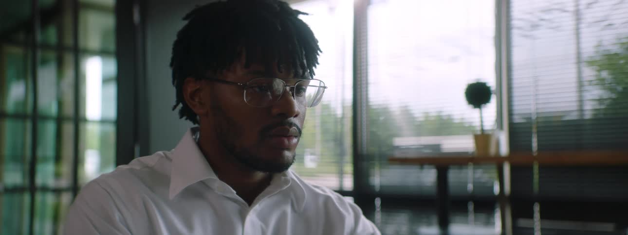 Man with braids glasses and a white shirt sits at a table and uses a grey laptop