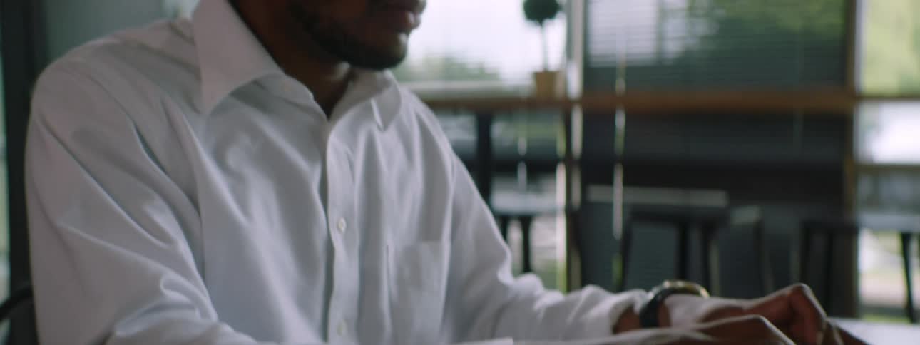 Black man with braided hair and a white shirt sits at a table and uses a laptop in a room