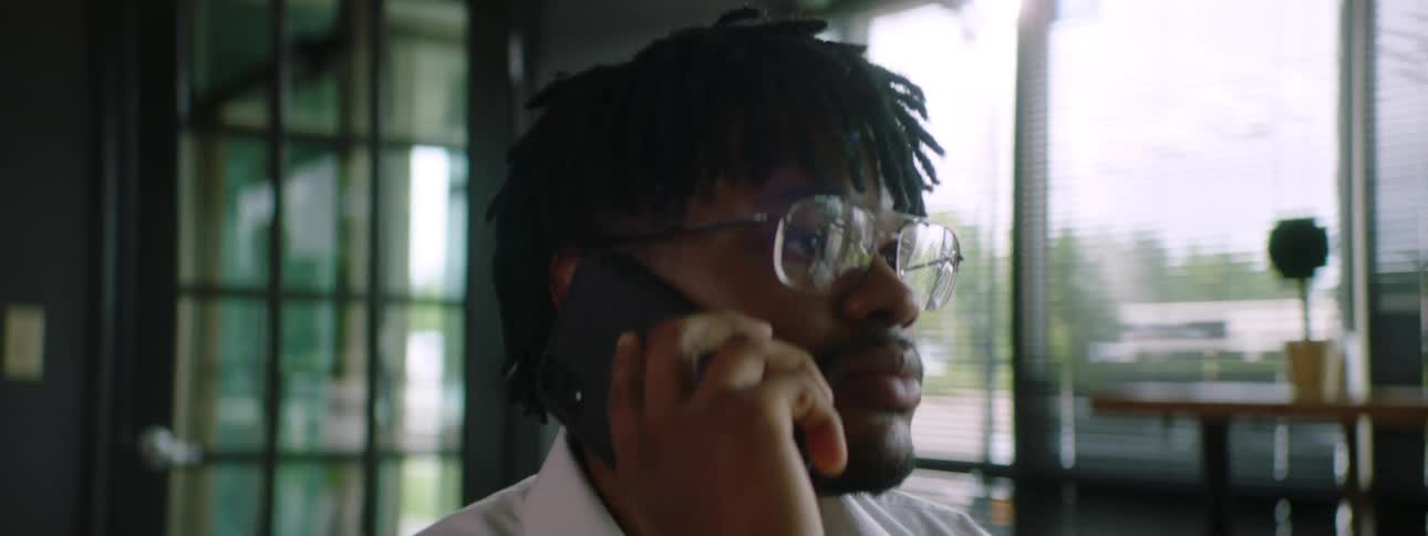 A black man sits in an office and talks on a cell phone