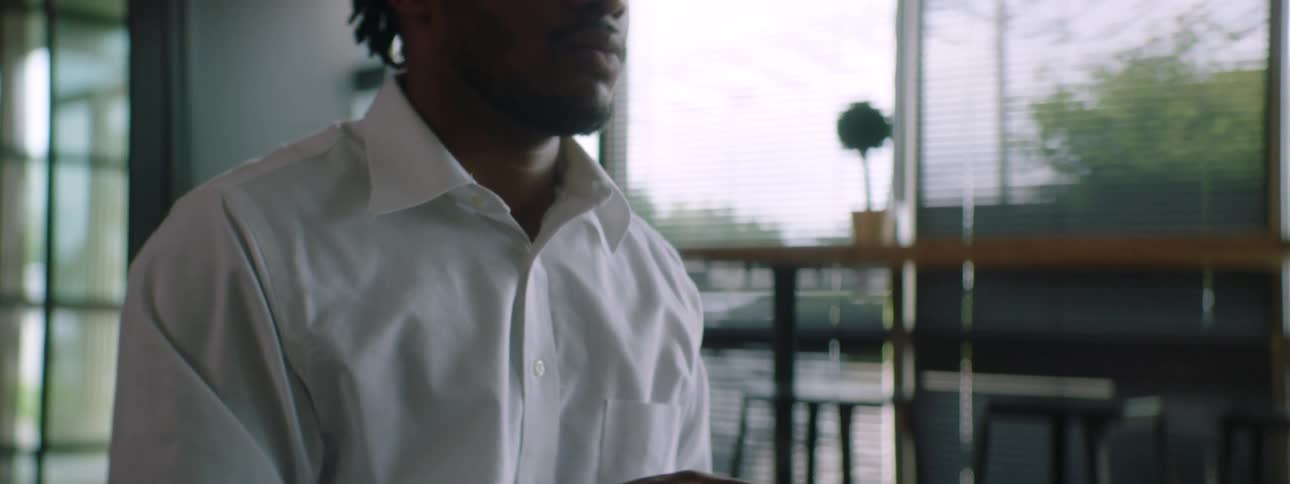 Black man with braids a beard and a white shirt sits at a table and uses a grey laptopthen thinks about something carefully in a room