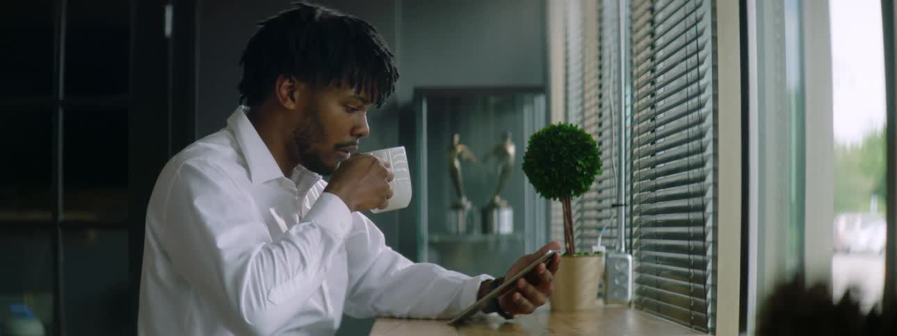 Black man with a braid hair a beard and a white shirt sits at a table and uses a smartphone while drinking a cup of coffee