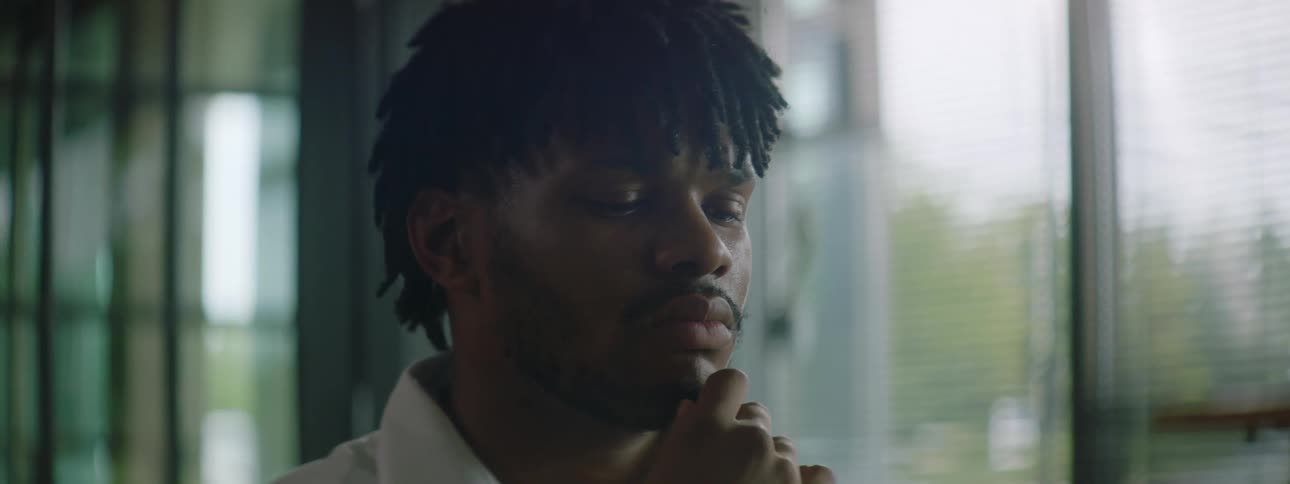 A man at home rubs his chin and looks out the window as he thinks