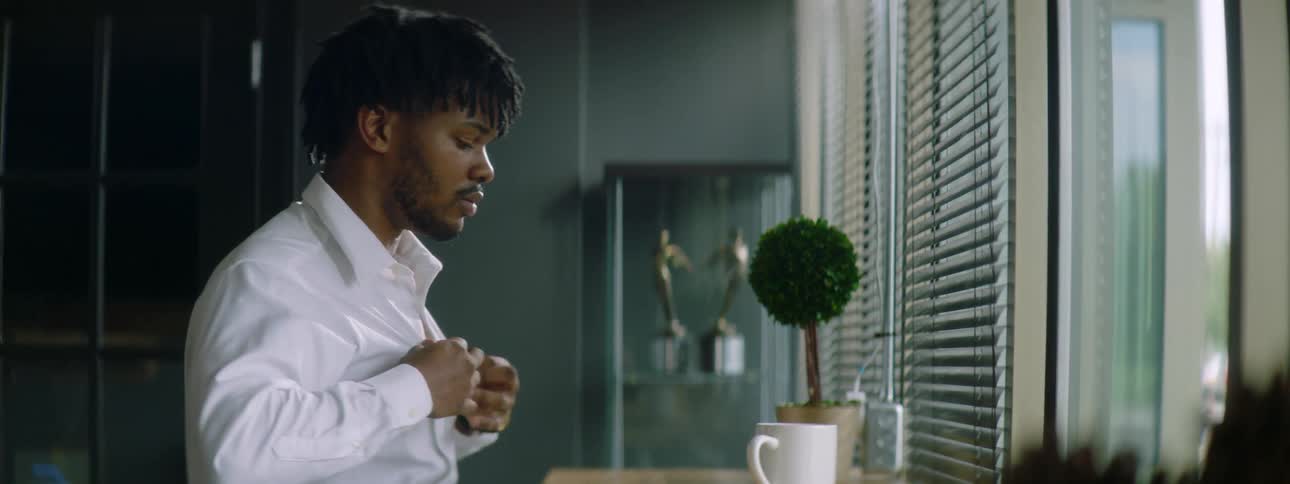 A young man places a tablet and coffee mug on a desk next to a window as he takes a seat before it