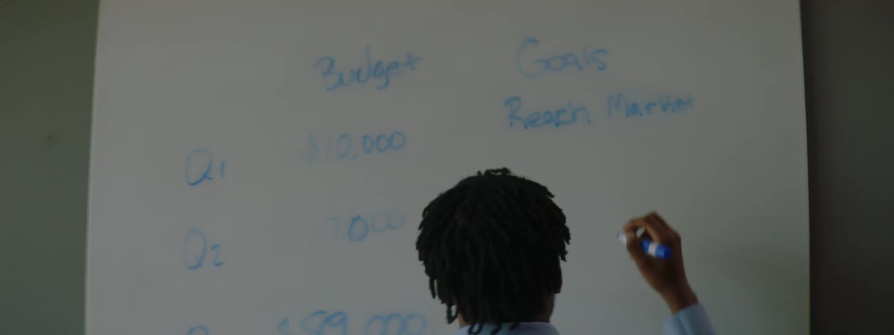 A man writing on a whiteboard with a marker stops to look down at his smartphone
