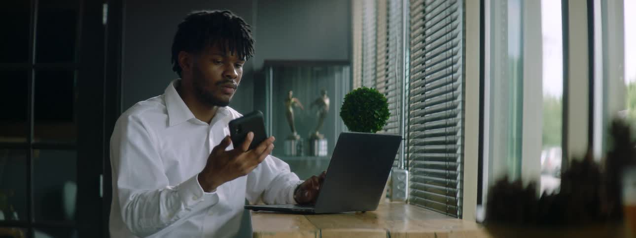 A man sitting at a desk while typing on a laptop stops to answer a call on his smartphone