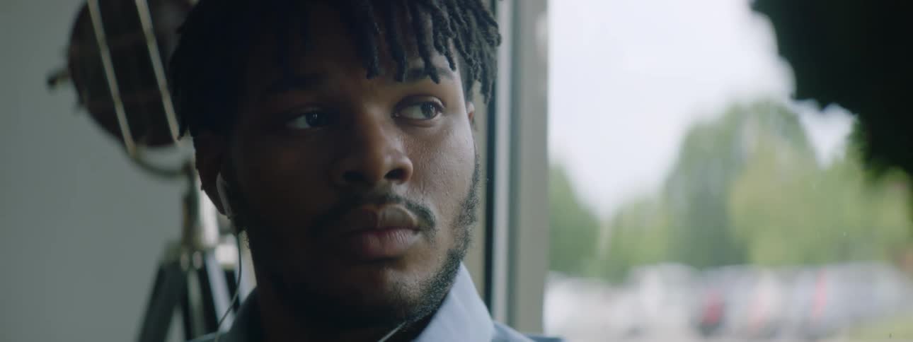 Black man with braids a beard and a blue shirt looks out the window while listening to music through earphones