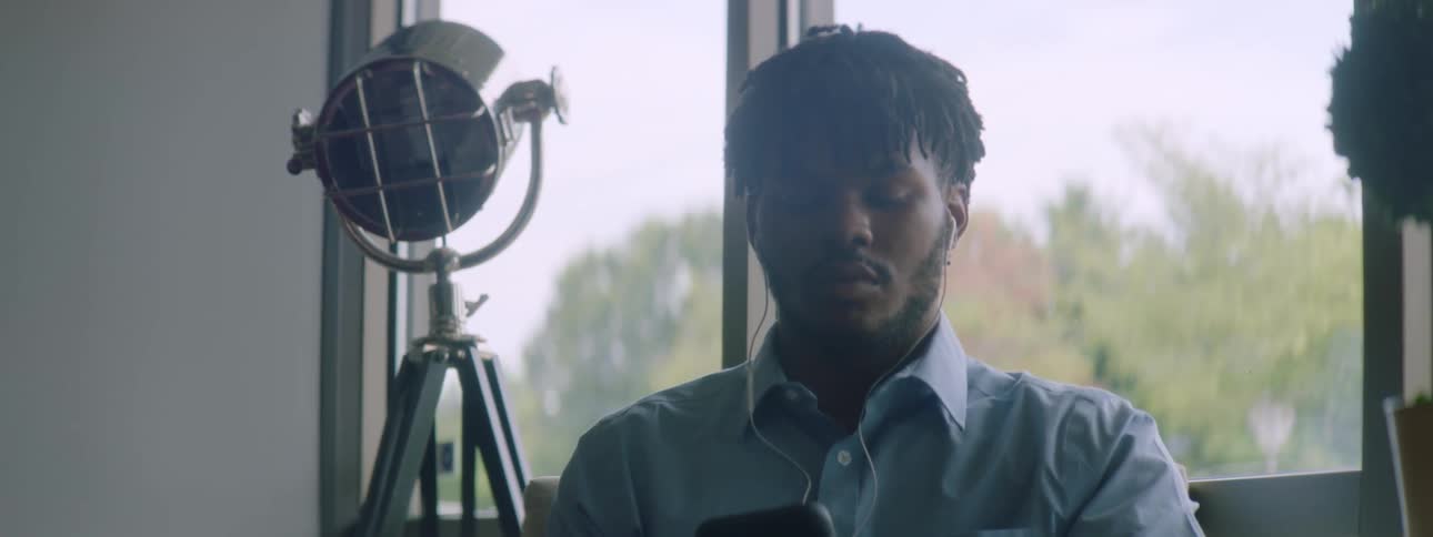 Black man with braided hair a beard and a blue shirt sits at a table and uses a smartphone with earphones
