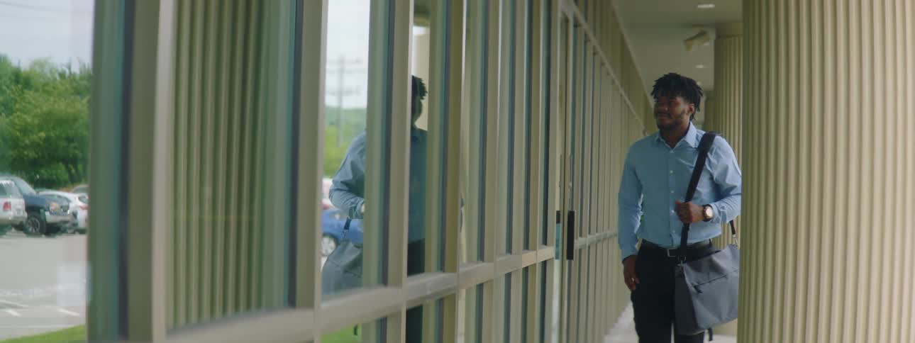 A young man carrying a bag walks along the outside of a building towards the camera