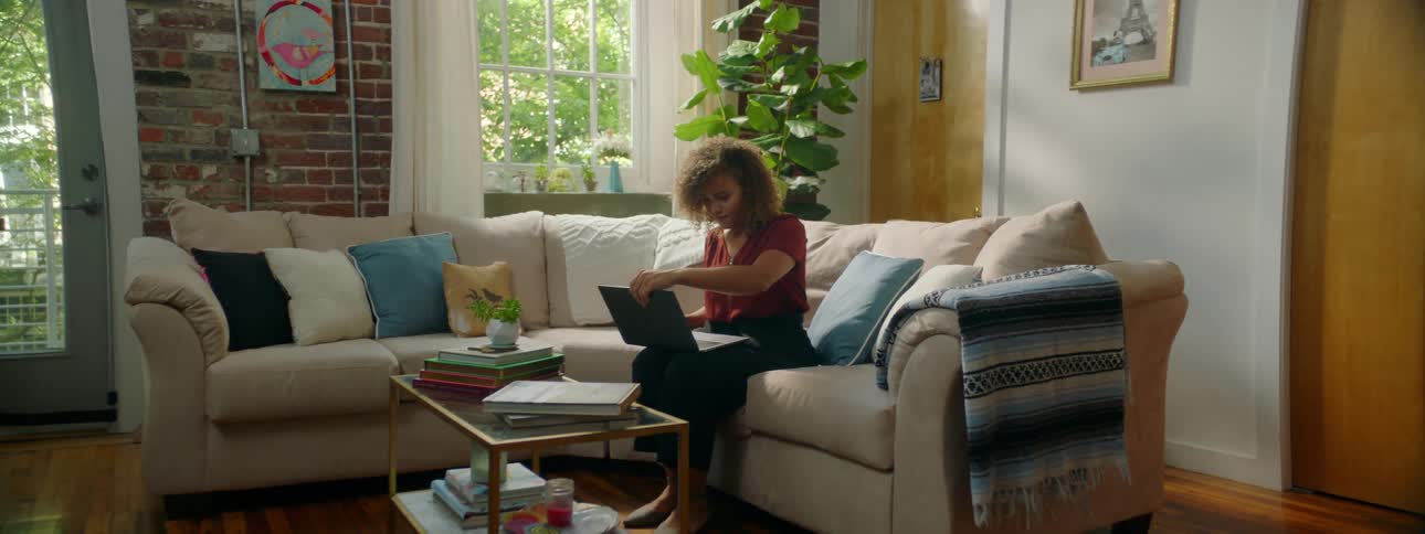 Woman with red curly hair and a red blouse sits on a sofa and opens her laptop in the living room at home