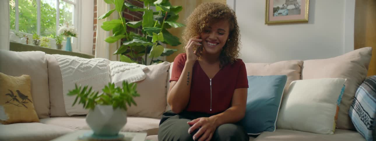 Female sitting on sofa at home laughs and smiles as she talks on her cell phone