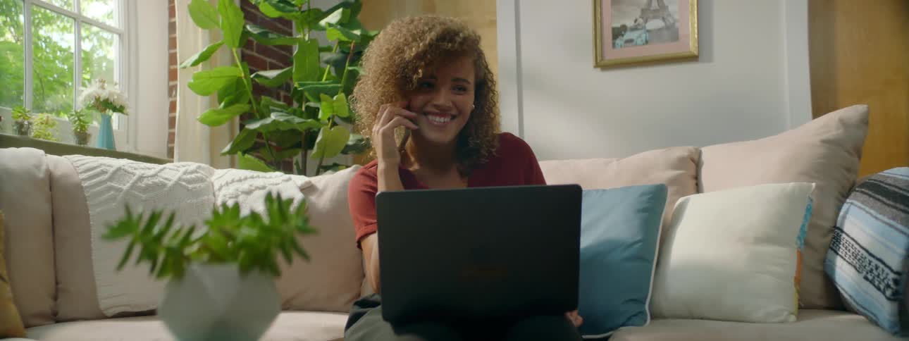 A smiling young woman talks on a smartphone while sitting on a sofa with her laptop