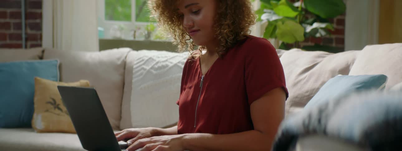 A woman is sitting on a sofa while typing on a laptop computer during the day