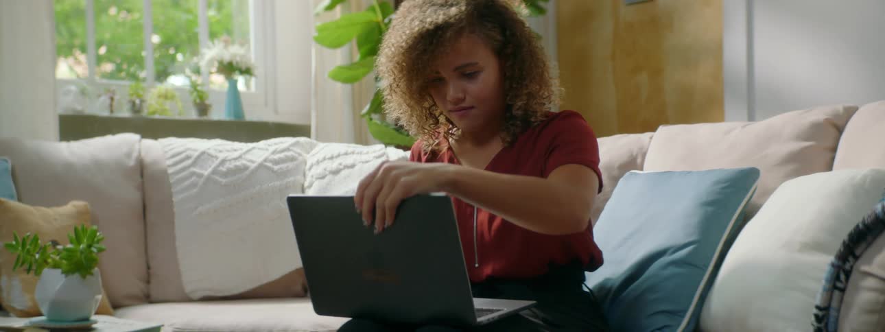 Woman with red curly hair and a red blouse sits on a sofa and opens a grey laptop in the living room at home