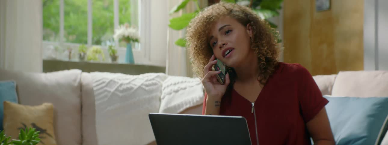 Woman with red curly hair and a blouse picks up a smartphone and answer the phone while using a laptop in the living room at home