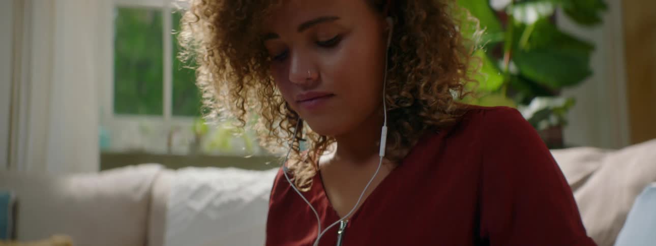 Woman with curly hair and a red blouse uses a smartphone and listens to music through earphones in the living room at home
