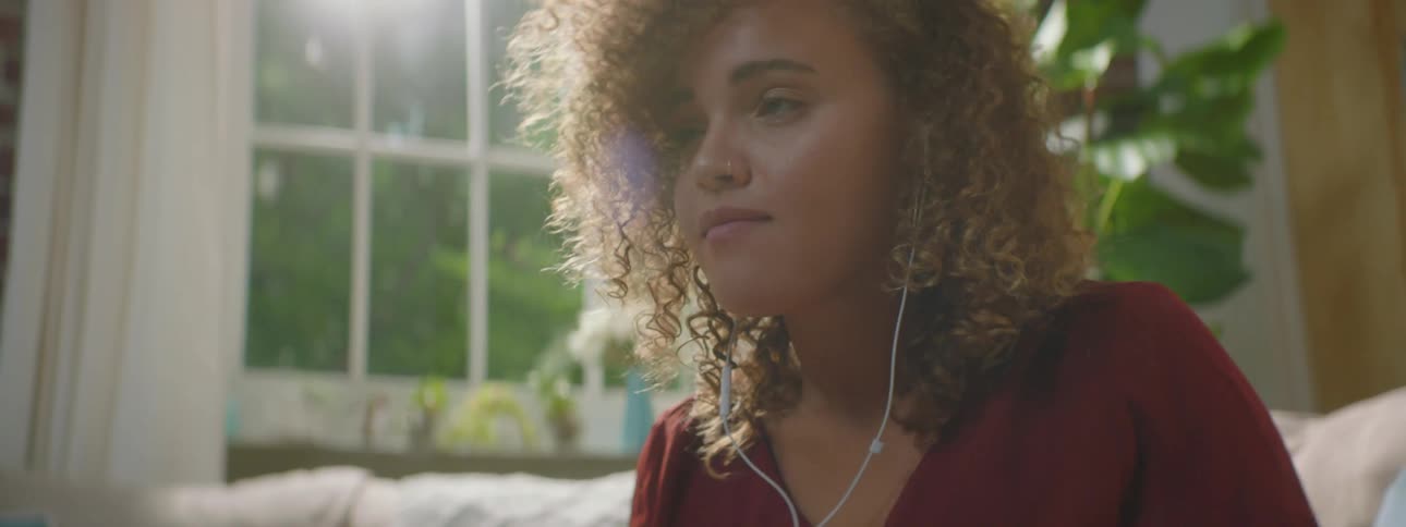 Woman with red curly hair and a red blouse sits on a beige sofa uses a smartphone and listens to music through earphones in the living room