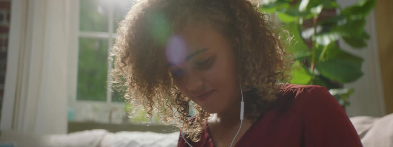 Woman with red curly hair and a red blouse uses a smartphone with earphones in the living room at home