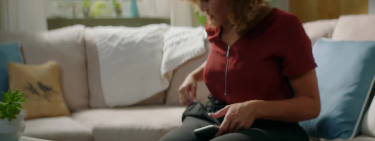 A young woman sitting on a sofa retrieves her smartphone from a table and a pair of tangled earbuds from her pocket