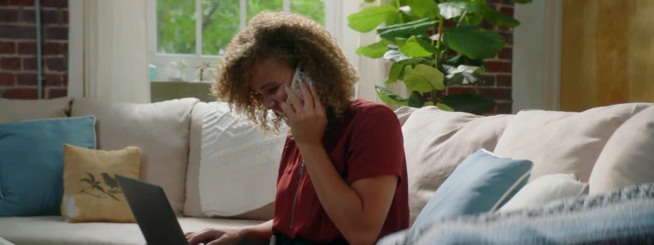 Woman sitting in the couch at home works on a laptop while talking on a smartphone