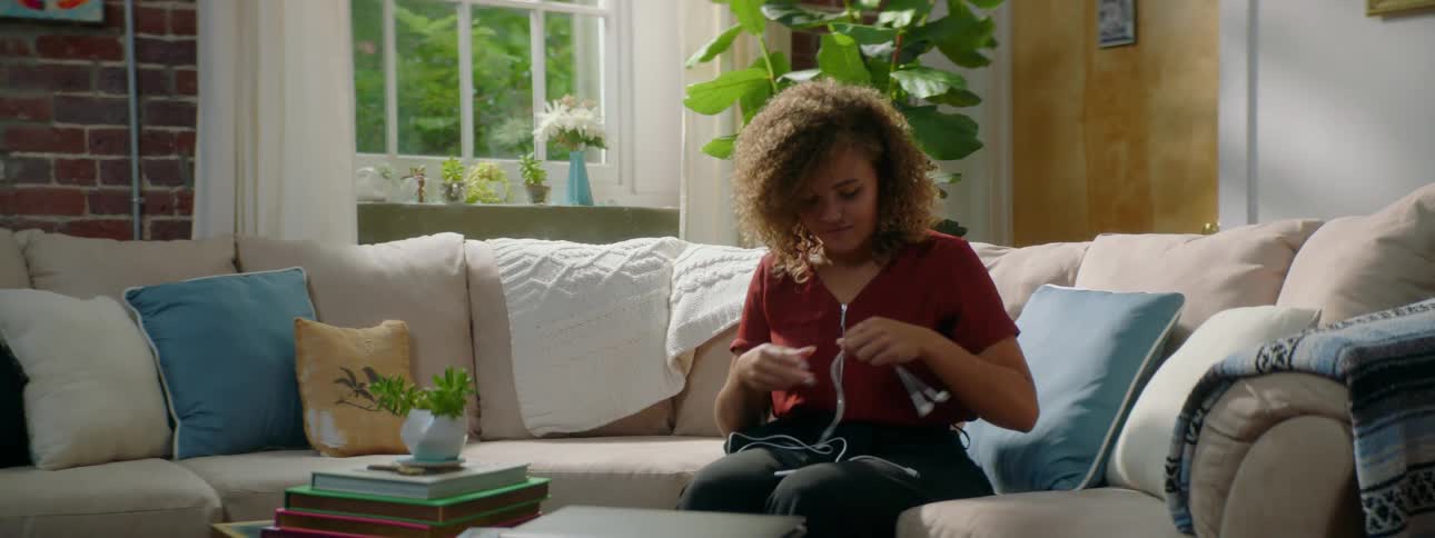 Young woman connects earbuds to her smartphone while sitting in a couch at home