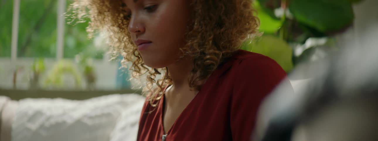 Woman with red curly hair and a red blouse sitting on a couch and types on a laptop and looks at the camera smilingly in the living room at home