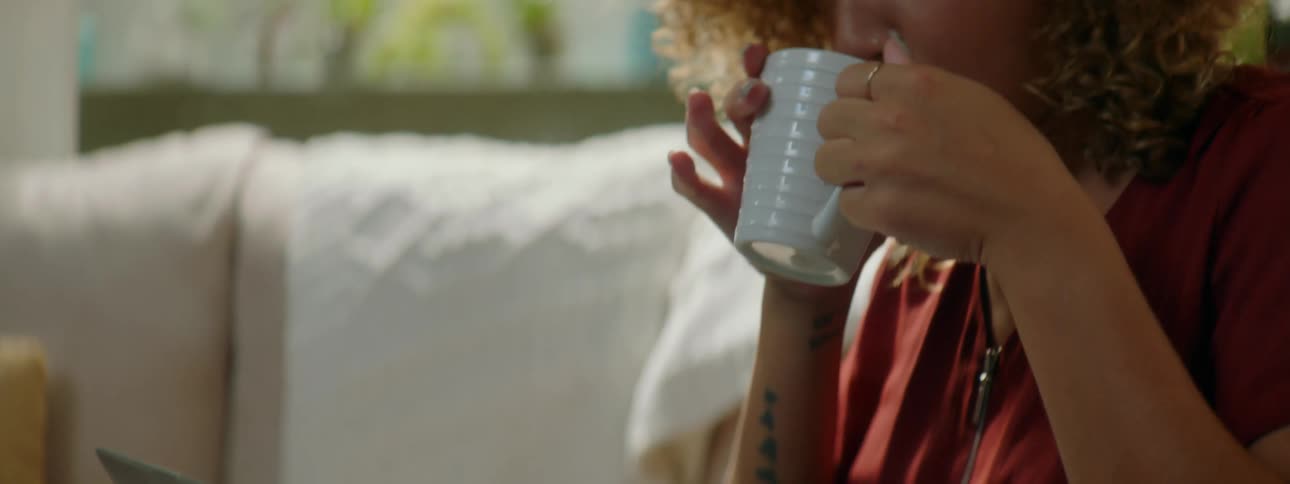 Woman with red curly hair and a red blouse sits on a sofa and drinks a cup of coffee while using a laptop in the living room at home