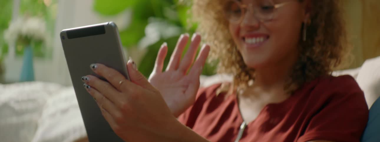 Woman with red curly hair glasses and a red blouse sits on a sofa and makes a video call on a grey tablet in the living room at home