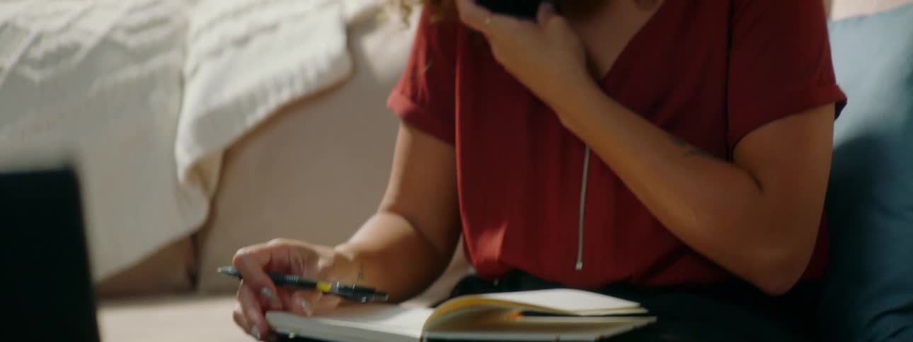 Woman with red curly hair a red blouse and glasses talks on the smartphone smilingly while writing in a notebook in the living room at home