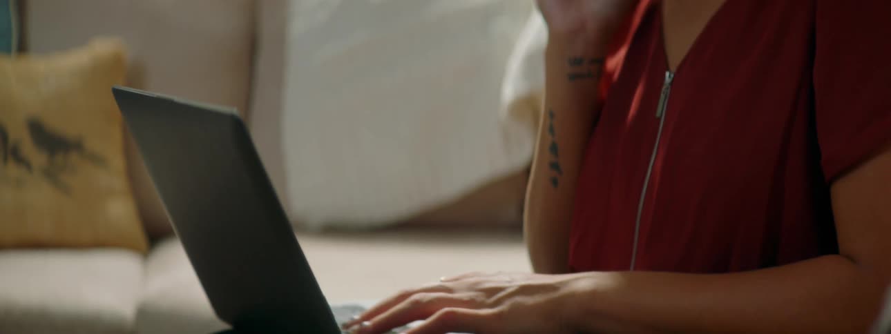 A young woman types on a laptop with one hand as she talks on her smartphone with the other while sitting on a sofa in the living room of a house
