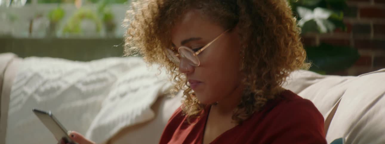 Woman with red curly hair and a red shirt sits on a sofa and uses a tablet in the living room
