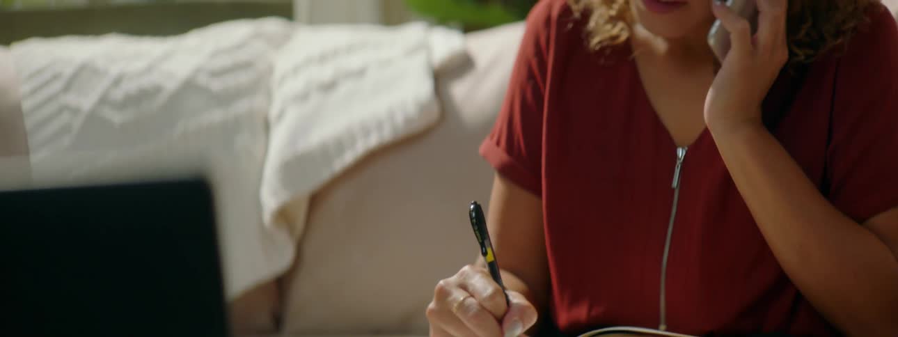 Woman with curly hair glasses and a red blouse writes in a notebook while talking on a smartphone in the living room at home