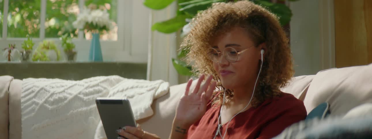 A woman sits on her living room couch holding a tablet and waves at someone during a video call