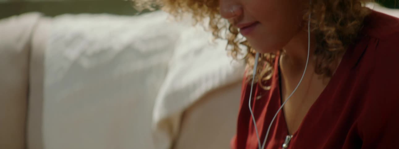 Woman with red curly hair and a red blouse uses a smartphone and listens to music through earphones in the living room at home