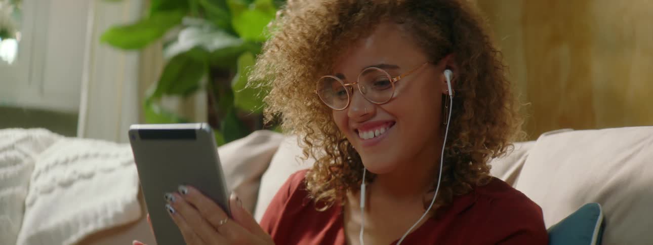 A teenage girl at home laughs and waves at her tablet as she video chats