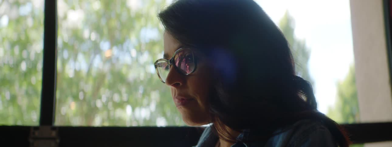 A woman uses a laptop while sitting at a table beside a large window during the day