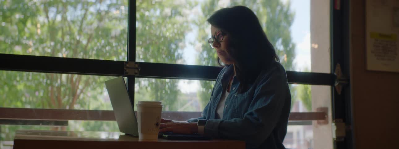 Woman with wavy hair a blue shirt and glasses sits at a table and uses a laptop in a cafe