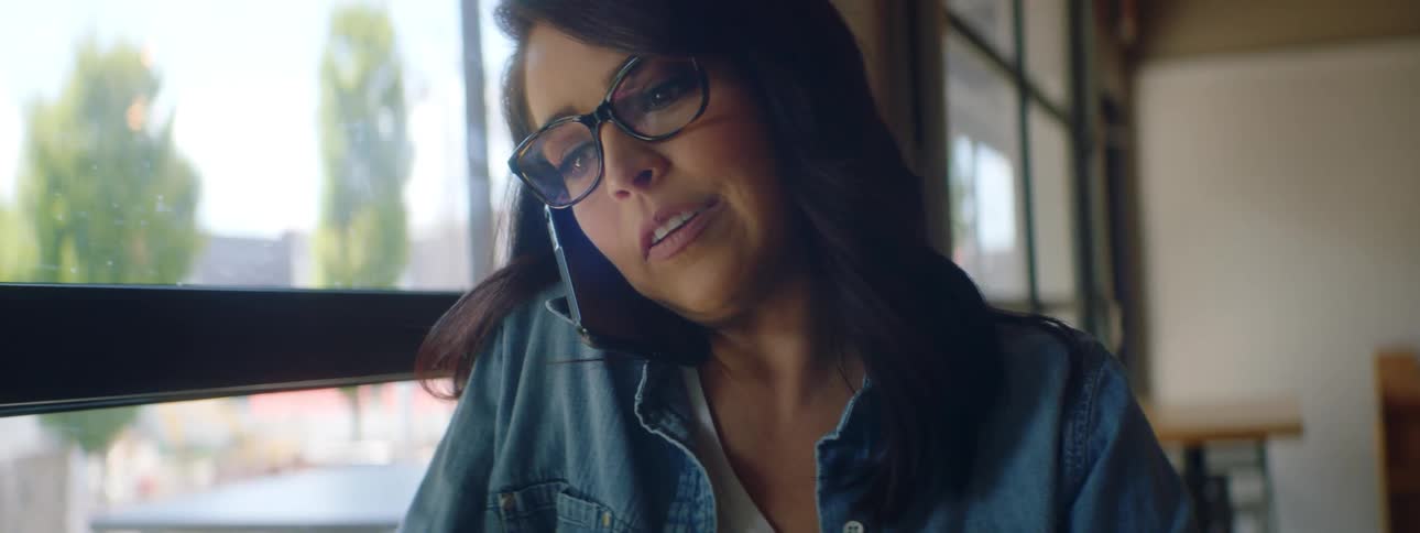Woman with a blue shirt and glasses sits at a table and answers the smartphone in a cafe
