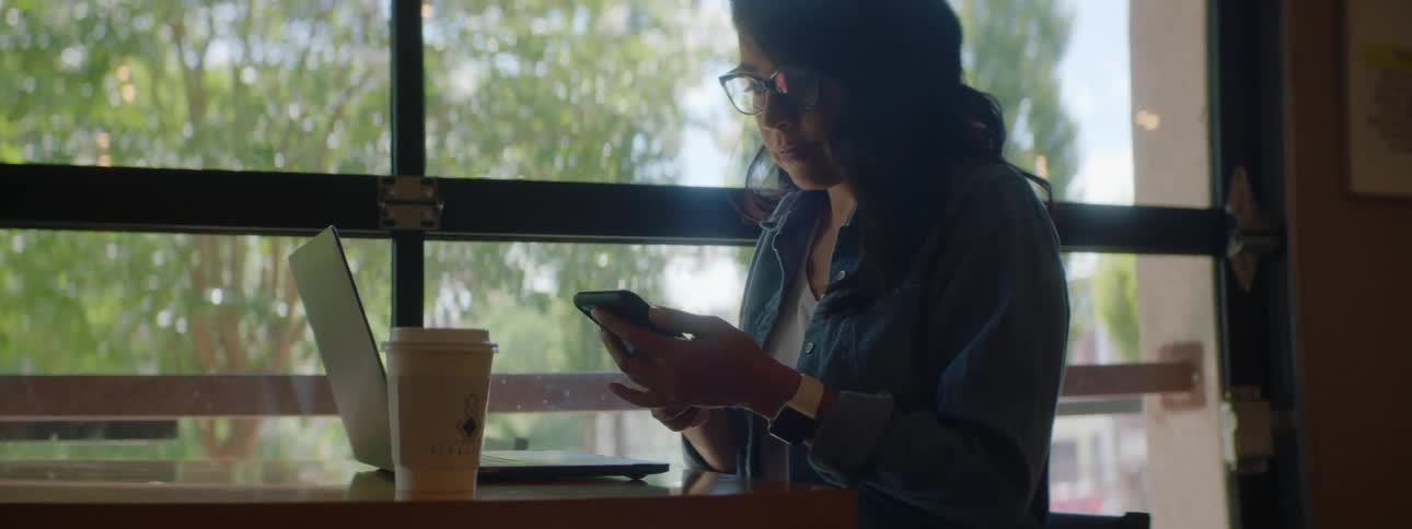 Woman with wavy hair and glasses answers the phone while using a laptop at a cafe