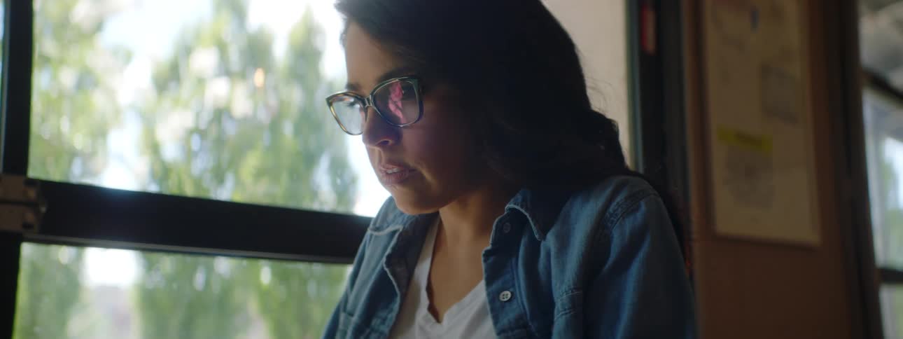 Woman with glasses and a blue shirt sits at a table and uses a grey laptop in a cafe