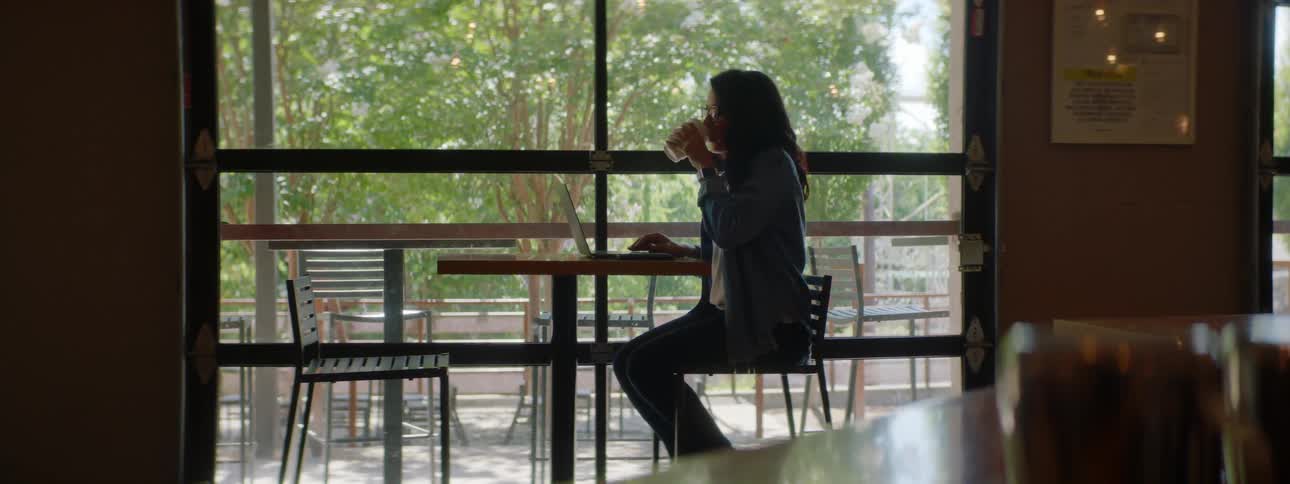 A woman sitting at a table typing on a laptop with another woman bringing a cup of coffee