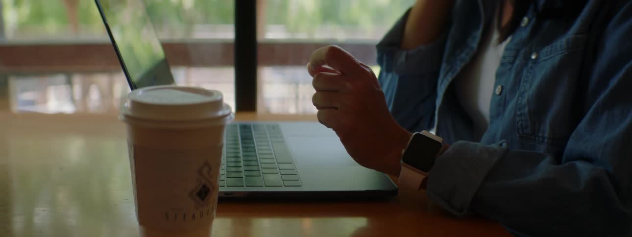 A young woman types on a laptop as she talks on her smartphone while sitting at a table indoors during the day